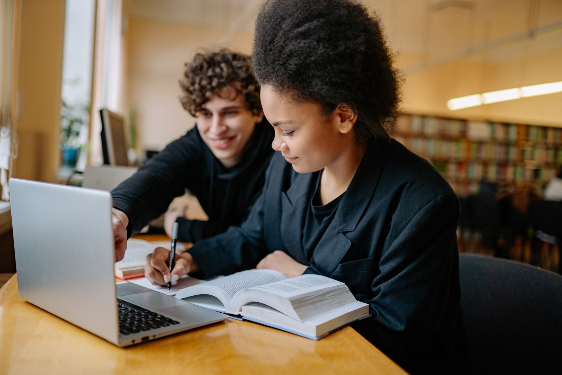 Two young adults studying together in a library, using a laptop and taking notes.